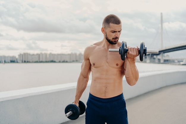 Un mec musclé avec des exercices de torse nu avec des haltères, se tient torse nu, entraîne les muscles en plein air, se tient debout sur piere, a la motivation de rester en forme et en bonne santé. Fitness, sport, concept de musculation