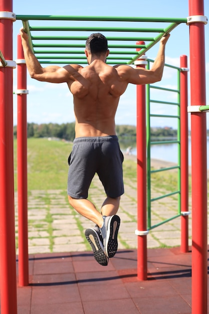 Mec musclé avec torse nu tirant sur une barre horizontale en plein air.