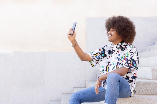 Mec noir aux cheveux afro souriant assis sur des marches de pierre prenant un selfie avec son smartphone