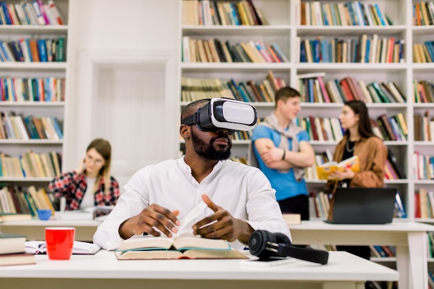 Mec noir portant un casque de lunettes vr, lisant un livre et utilisant des informations de la réalité virtuelle, assis dans une bibliothèque moderne. Groupe d'étudiants qui étudient et se parlent