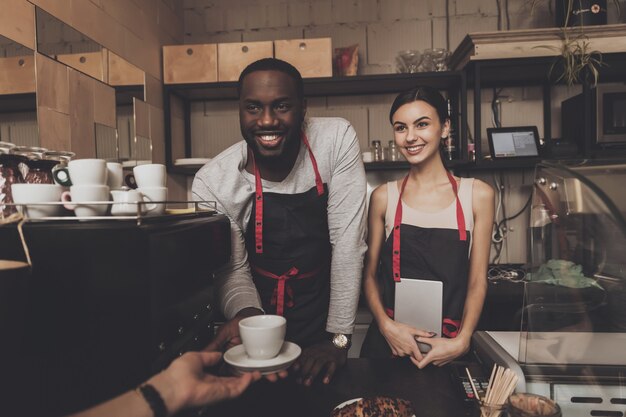 Un mec noir souriant en tablier donne une tasse de café cuit au visiteur dans un café. Confiserie. Barista.