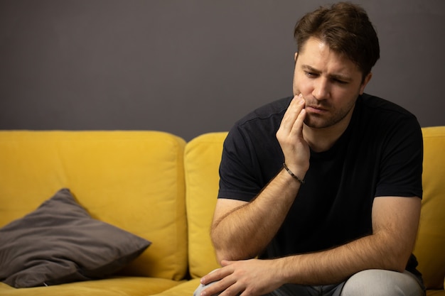 Un mec non rasé est assis sur un canapé et souffre de maux de dents dentiste aide les soins dentaires