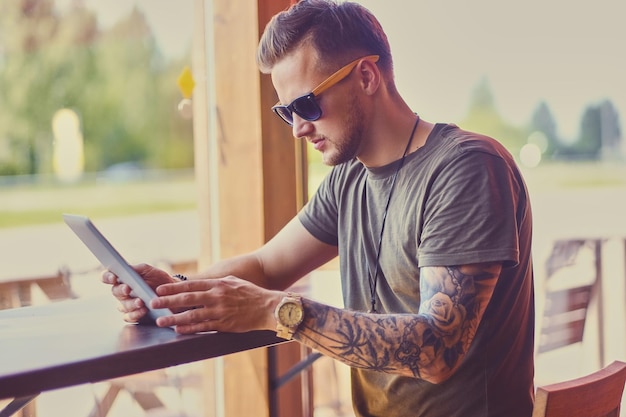 Un mec séduisant en lunettes de soleil à l'aide d'une tablette PC dans un café près de la fenêtre.