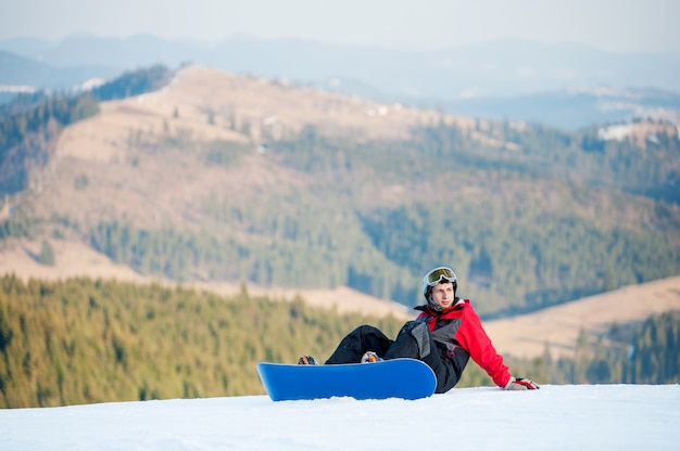 Mec avec snowboard assis sur une pente enneigée au sommet d&#39;une montagne et regardant ailleurs