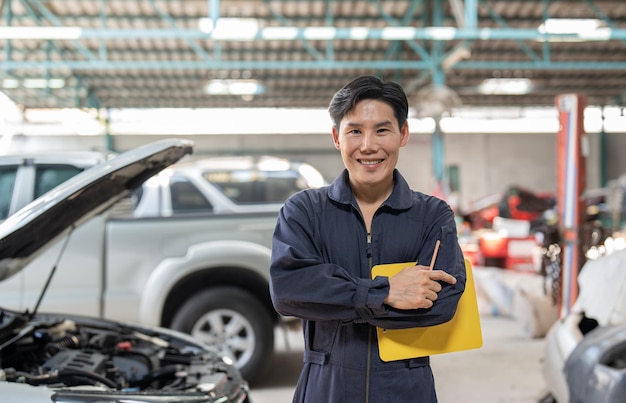 Photo un mécanicien asiatique souriant avec un bloc-notes debout les bras croisés au service de garage automobile