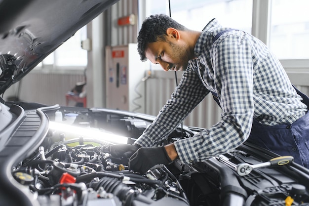 Mécanicien automobile indien debout et travaillant dans une station-service Spécialistes de l'automobile examinant la voiture levée Réparateurs professionnels portant l'uniforme de mécanicien de couleur bleue