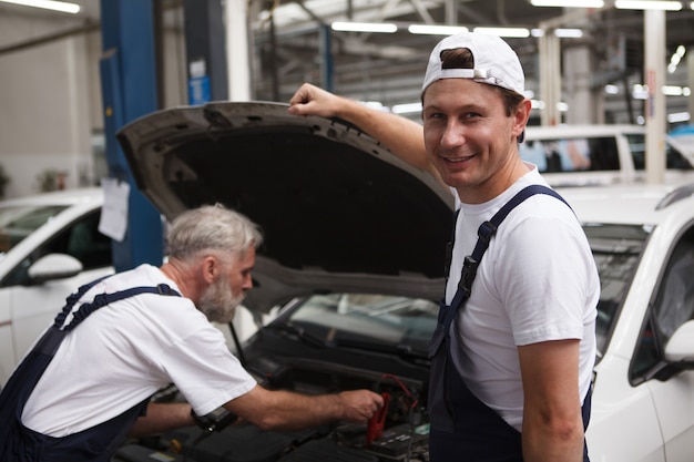 Mécanicien automobile joyeux souriant à la caméra tout en travaillant sur une voiture avec son collègue senior