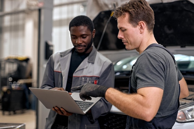 Un mécanicien automobile noir et un réparateur caucasien vérifiant les résultats des diagnostics sur un ordinateur portable expliquent...