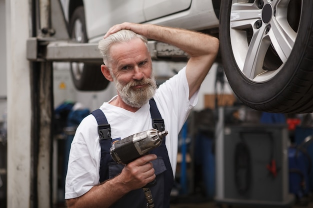 Mécanicien automobile senior confiant souriant à la caméra, travaillant avec une clé dynamométrique électrique dans son atelier