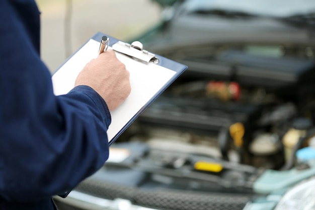 Photo mécanicien debout devant un capot de voiture ouvert avec presse-papiers et stylo gros plan