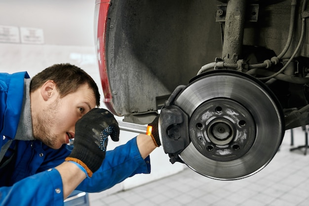 Mécanicien masculin avec la bouche ouverte portant des gants noirs et uniforme bleu, tenant une lampe de poche et examinant soigneusement les pneus ou les plaquettes de frein de voiture levée à l'atelier automobile Concept de service et de technicien de voiture