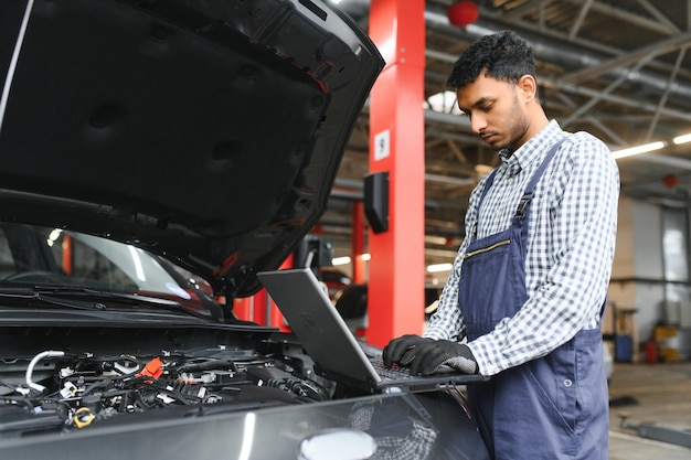 Mécanicien mécanicien ouvrier à l'aide d'un ordinateur portable vérifiant la voiture dans l'atelier du centre de service de réparation de voitures automobiles Ingénieur jeune homme regardant les détails du véhicule d'inspection sous le capot de la voiture