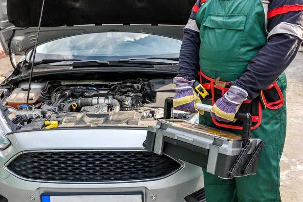 Mécanicien en uniforme posant avec une boîte à outils près du moteur de la voiture