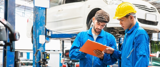 Un mécanicien en uniforme de vêtements de travail bleu vérifie la liste de contrôle d'entretien du véhicule avec son assistant avec une voiture levée floue en arrière-plan Service de réparation automobile Travail d'équipe professionnel