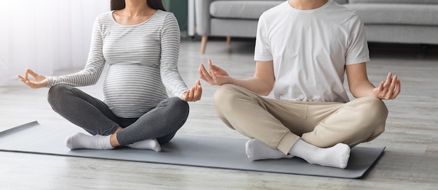 Méconnaissable attendant une famille méditant ensemble, recadrée d'un jeune homme et d'une femme enceinte ayant un mode de vie sain pendant la grossesse, assis sur un tapis de fitness et pratiquant le yoga à la maison, panorama