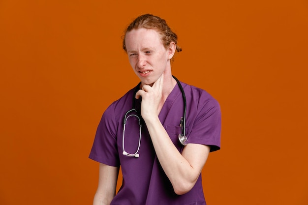 Mécontent de mettre la main sur le cou jeune homme médecin portant l'uniforme avec stéthoscope isolé sur fond orange