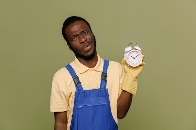 Mécontent tenant réveil jeune homme nettoyeur afro-américain en uniforme avec des gants isolés sur fond vert