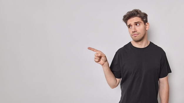 Le mécontentement bouleversé L'homme adulte européen pointe avec l'index montre quelque chose de désagréable qui n'aime pas cette publicité vêtue d'un t-shirt noir isolé sur fond blanc. Espace maquette