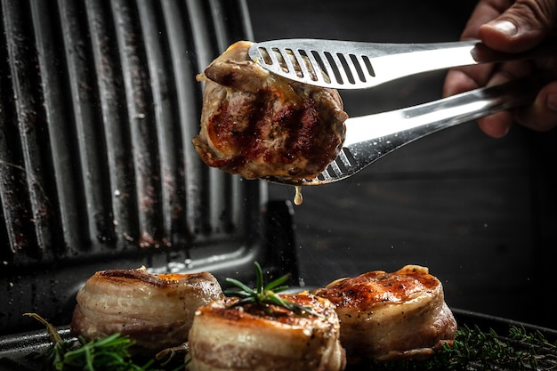 Médaillons de steaks du filet de bœuf recouvert de bacon sur le gril Fond sombre. Cuisson du steak de boeuf sur le gril par les mains du chef.