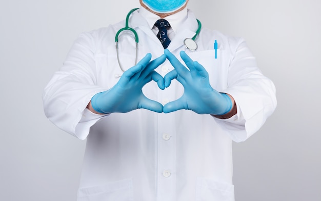 Médecin adulte homme dans une blouse médicale blanche avec un stéthoscope sur le cou montre un geste cardiaque avec ses mains, portant des gants bleus