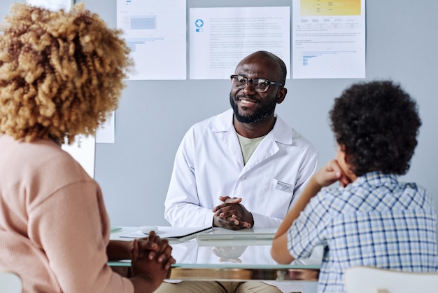 Médecin africain assis à sa table et discutant avec une femme de la maladie de son fils