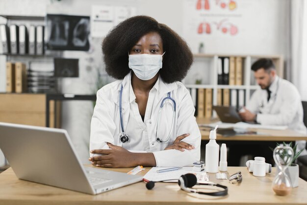 Médecin africain en masque facial assis au bureau avec ordinateur portable