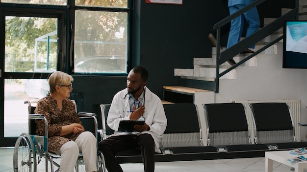 Médecin afro-américain consultant une vieille femme en fauteuil roulant, parlant de handicap physique et de maladie dans la salle d'attente de l'hôpital. Spécialiste et patient handicapé faisant un examen médical.