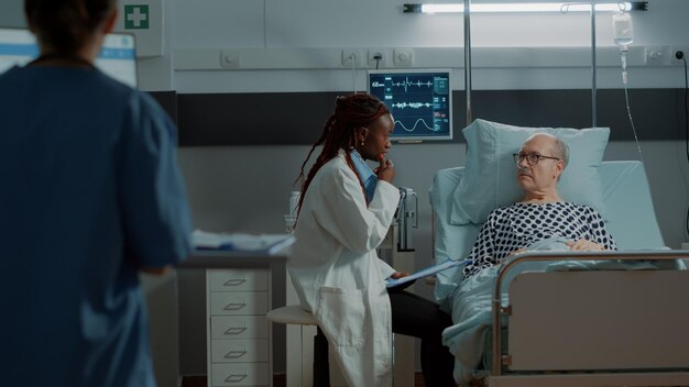 Un médecin afro-américain consulte un patient âgé malade dans une salle d'hôpital pendant que l'infirmière rédige une ordonnance sur ordinateur. Vieil homme avec oxymètre et tube à oxygène nasal recevant un traitement