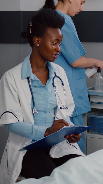 Médecin afro-américain discutant avec un homme malade écrivant un traitement médical sur le presse-papiers dans la salle d'hôpital pendant la réadaptation de la maladie