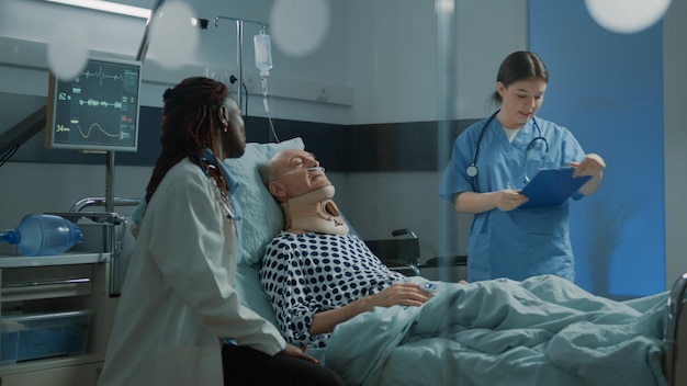 Un médecin afro-américain parle à un patient malade dans une salle d'hôpital et une infirmière vérifie les symptômes. Vieil homme avec collier cervical assis dans son lit en salle de soins intensifs pour guérir les problèmes