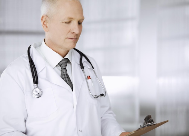 Un médecin âgé et confiant se tient droit et utilise le bloc-notes dans la clinique. Un service médical parfait. Le concept de médecine et de soins de santé.