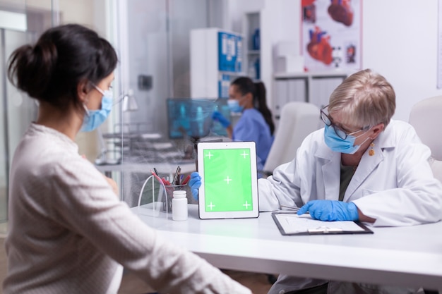 Médecin âgé tenant une tablette avec écran vert dans la salle de la clinique parlant avec un jeune patient expliquant le traitement en regardant l'affichage de la maquette. Affichage au moniteur izolated, bureau d'incrustation de chrominance.