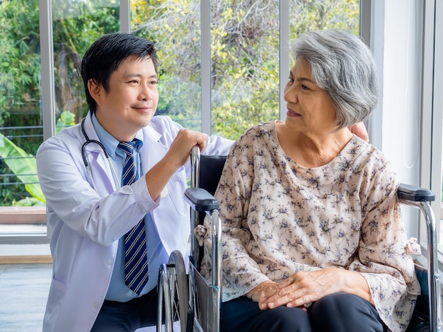 Un médecin asiatique positif souriant en blouse blanche parle avec soin à une patiente âgée âgée qui est assise dans un fauteuil roulant dans un cabinet médical à l'hôpital Soins de santé et concept médical