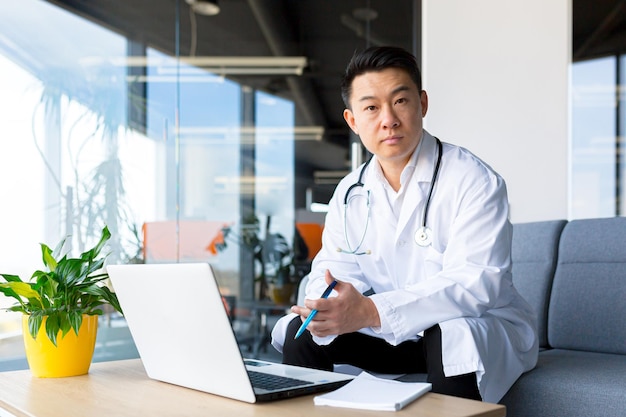 Médecin asiatique prospère travaillant sur un ordinateur portable dans une clinique moderne