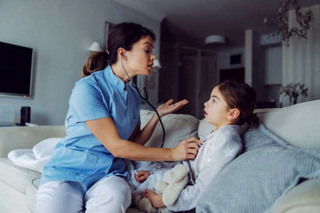 Médecin assis sur le canapé à côté de la fille et examinant ses poumons avec un stéthoscope. Médecin au service à domicile.