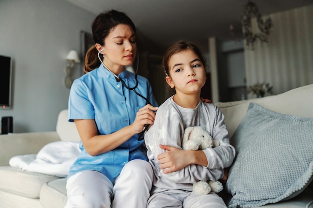 Médecin assis sur un canapé à côté d'une fille et examinant ses poumons avec un stéthoscope