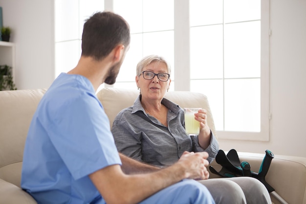Médecin assis sur un canapé dans une maison de retraite parlant avec une femme âgée avec des béquilles pendant qu'elle prend ses pilules.