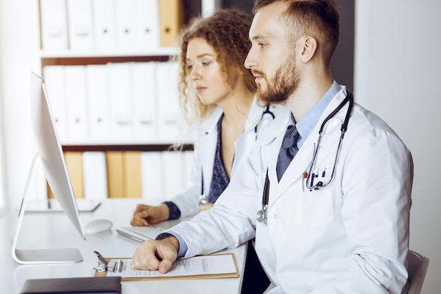 Médecin à barbe rouge avec une collègue discutant de la thérapie actuelle de la maladie tout en étant assis sur le lieu de travail dans une clinique ensoleillée. Travail d'équipe en médecine.