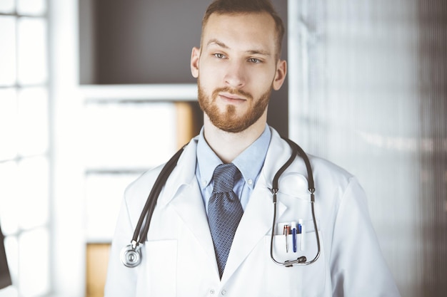 Médecin à barbe rouge debout directement dans une clinique près de son lieu de travail. Portrait de médecin. Concept de médecine et de soins de santé.