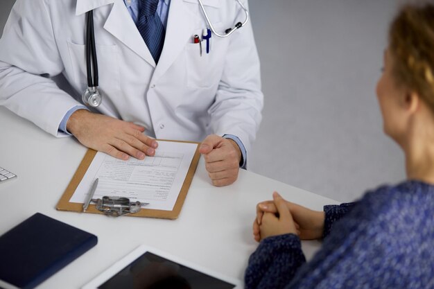 Médecin barbu inconnu et femme patiente discutant de l'examen de santé actuel tout en étant assis dans une clinique ensoleillée, en gros plan. Notion de médecine