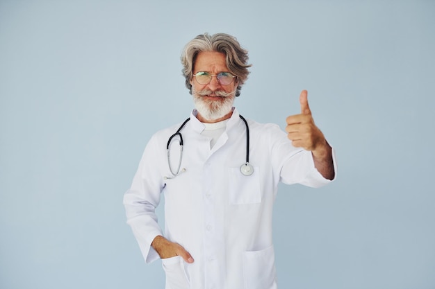 Médecin en blouse blanche contre le mur Senior homme moderne et élégant aux cheveux gris et à la barbe à l'intérieur