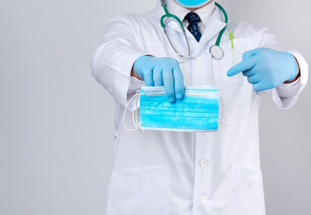 Médecin en blouse blanche, gants stériles en latex bleu, tenant des masques médicaux en textile à la main, accessoire de protection contre les virus