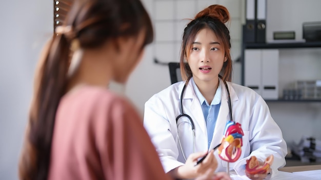 Photo médecin cardiologue asiatique femmes pointant sur le modèle anatomique du cœur humain pour expliquer les données médicales et donner des conseils de thérapie de santé mentale à une patiente après avoir examiné la santé en clinique