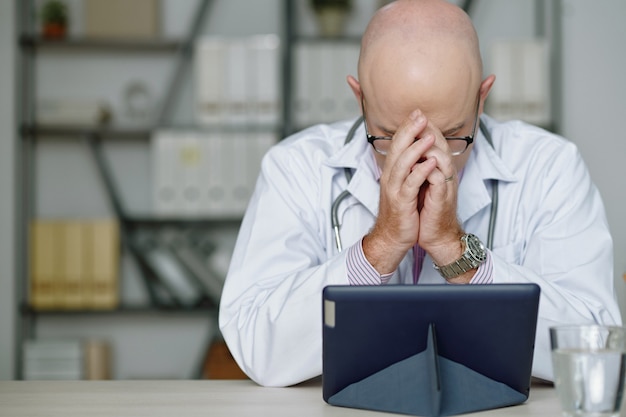 Un médecin caucasien chauve perplexe en blouse blanche fatigué du travail assis à table et couvrant le visage avec les mains tout en lisant des informations sur une tablette au bureau