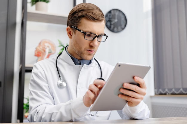 Médecin caucasien masculin en uniforme médical blanc remplissant les antécédents médicaux ou l'anamnèse du patient sur tablette au bureau Médecin consultant le client en ligne sur tablette dans le concept de soins de santé clinique moderne