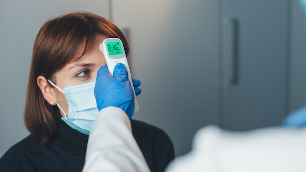 Un médecin caucasien portant des gants mesure la température corporelle du patient avec un masque lors d'une consultation