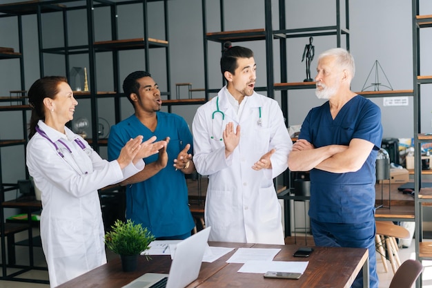 Le médecin-chef masculin mature aux cheveux gris et heureux qui réussit reçoit les applaudissements de l'équipe dans la salle de bureau lumineuse de l'hôpital moderne. Concept de médecine et de soins de santé.