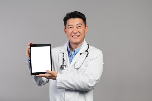 Médecin chinois mature souriant en blouse blanche avec stéthoscope montrant une tablette avec écran vide