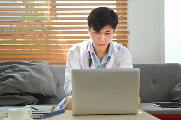 Médecin concentré en blouse blanche assis au bureau et regardant une formation en ligne sur un séminaire médical en ligne sur un ordinateur portable