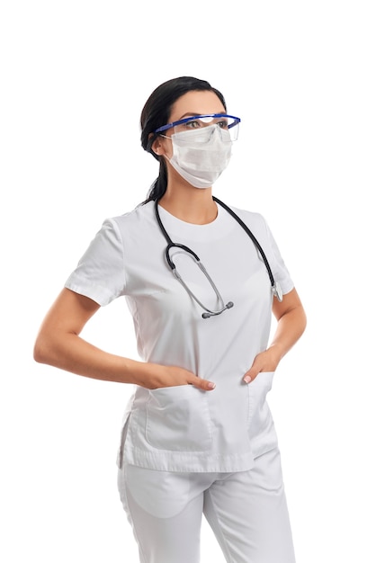 Médecin confiant dans un masque de protection et des lunettes se tenant la main dans des poches d'uniforme médical